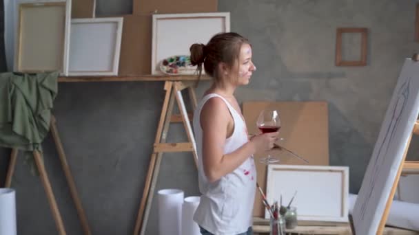 One woman artist with a glass of red wine and a palette of paints approaches an unfinished painting, drinks a drink and looks. — Video