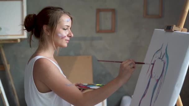 A young woman in a white dirty T-shirt stands in an art studio and paints a picture of a womans silhouette with a brush. — Video