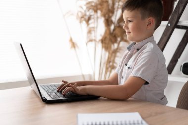 Side view of a schoolboy at home schooled working on a laptop typing text on the keyboard while communicating with a remote teacher. The child is studying computer programs online. Education concept.