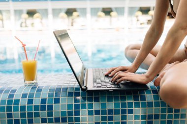 hands of a woman manager typing text on a laptop keyboard sitting by a swimming pool with clear water at a resort on vacation. Vacation and remote work of an emigrant. Communication