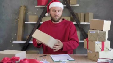 Delivery service for christmas and new year, young bearded Caucasian male businessman sitting at a table with boxes preparing them for delivery. Many more boxes are brought to him.