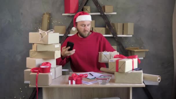 Christmas delivery service, a young bearded male businessman sits in a red sweater and santa claus hat takes a box scans a barcode using his smartphone. — Vídeo de Stock