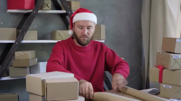 Kaukasischer bärtiger Mann mit Weihnachtsmannmütze und rotem Pullover, der zu Hause arbeitet. Nimmt Bastelpapier und packt die Schachtel. Konzept des Verkaufs von Waren für Weihnachten und Neujahr. — Stockvideo