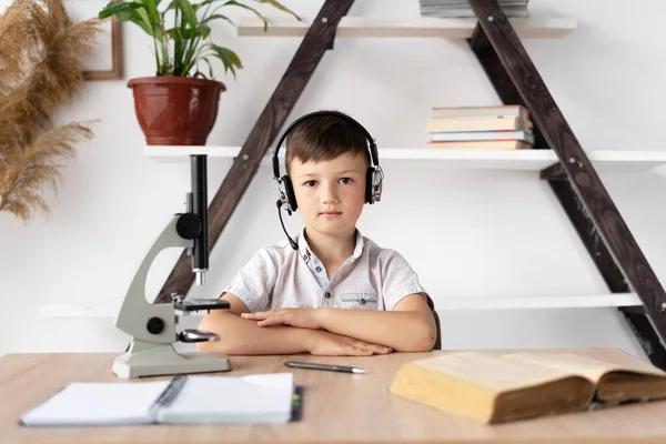 boy student in headphones is studying at home has a biology lesson and a microscope. Online education and home teacher. A student listens to a webinar via a wireless headset. Close-up.