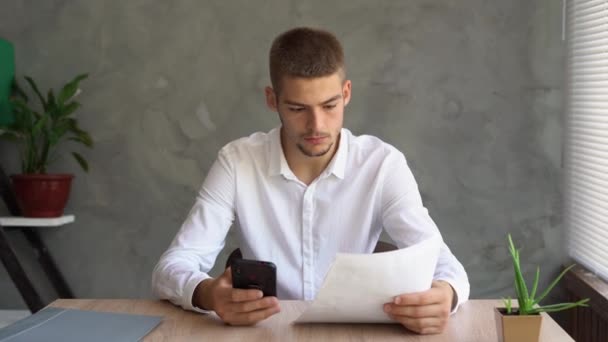 Συγκεντρωμένος σοβαρός καυκάσιος γενειοφόρος αναλυτής λογιστής επιχειρηματίας κρατώντας έγγραφα, κοιτάζοντας την οθόνη της έρευνας μάρκετινγκ smartphone του, σκέψης, εργασίας, κάθεται στο τραπέζι του γραφείου στο σπίτι. — Αρχείο Βίντεο