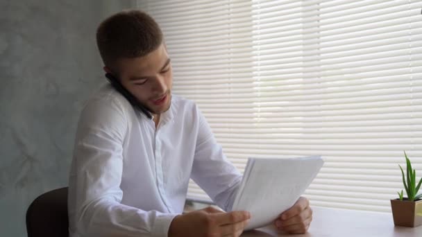Biały mężczyzna biznesmen siedzi w biurze przy stole trzymając papiery z wykresami w rękach i bawiąc się rozmawiając przez smartfona. Analityk koncepcyjny cieszy się z udanej transakcji. — Wideo stockowe