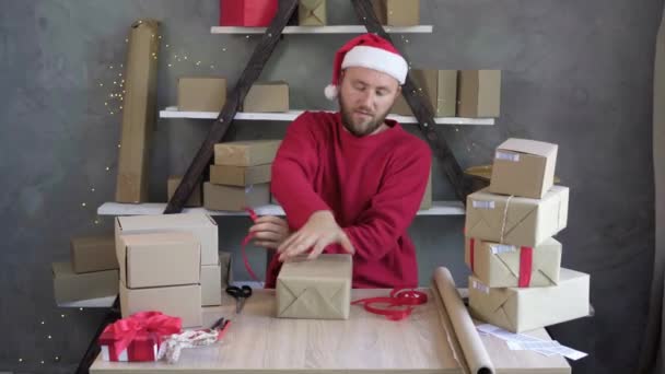 Noel Baba şapkası takan bir erkek işadamı kutu paketliyor ve kırmızı kurdele örüyor. Noel malları üzerine iş konsepti, bir sürü satış.. — Stok video