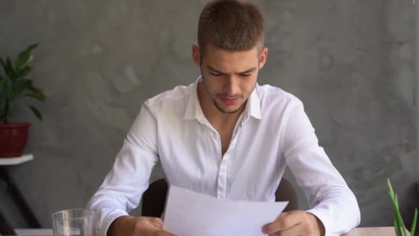 Um jovem caucasiano senta-se em uma mesa em casa em um escritório estudando dados em um papel, esmigalha uma folha e joga-a fora. conceito comerciante examina relatórios, mau negócio. — Vídeo de Stock