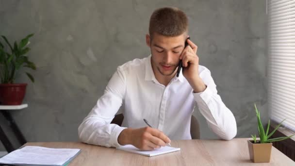 L'imprenditore caucasico seduto a casa in ufficio risponde alla telefonata e prende nota con una penna nel suo quaderno. Il concetto di vendite telefoniche, chiamate fredde. — Video Stock