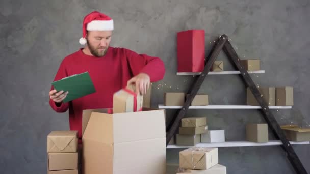 Un dueño de un negocio masculino está trabajando empacando una orden para ser enviada a un cliente. con un sombrero de Santa Claus y un suéter rojo. Hombre de negocios caja de embalaje para la entrega. — Vídeos de Stock