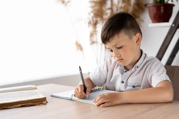 机の上でクラスに座りながらノートに手書きの宿題をする真剣な男子学生 テストのために小学生の男の子を準備する 家で教育や知識を得ること 家庭教育 — ストック写真