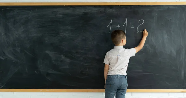 子供のバックビューは 数学の例の黒板のソリューションにチョークで書き込みます 小学校数学の授業 少年は黒板の近くで考えている 授業中の生徒 教育理念 — ストック写真