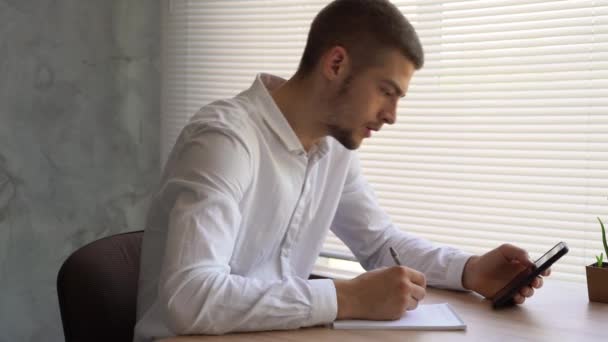 Um empresário masculino senta-se em seu escritório olhando para seu smartphone e faz anotações em seu caderno. conceito estudante procurando informações na internet. — Vídeo de Stock
