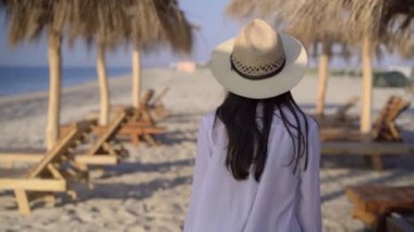 Genç, güzel, beyaz bir bayan, sahil boyunca saman şemsiyeler arasında tatil köyünde yürüyor. Kameraya bakar ve gülümser..