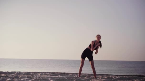 Egy fiatal nő bemelegít a parton. Naplemente és óceán a háttérben. Wellness és sportos életmód. — Stock videók