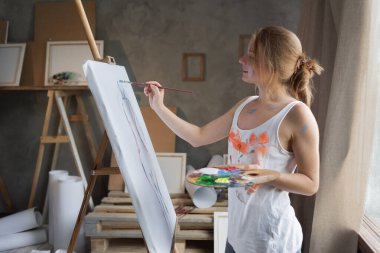 Kirli tişörtlü beyaz bir kadın ressam bir sehpanın yanında duruyor ve tuvale boya darbeleri uyguluyor. En sevdiğim yaratıcı hobi konsepti. Boşluğu kopyala.