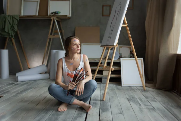 stock image a young beautiful woman artist sits on the floor in an art studio holding a paintbrush in her hands. favorite creative hobby concept.