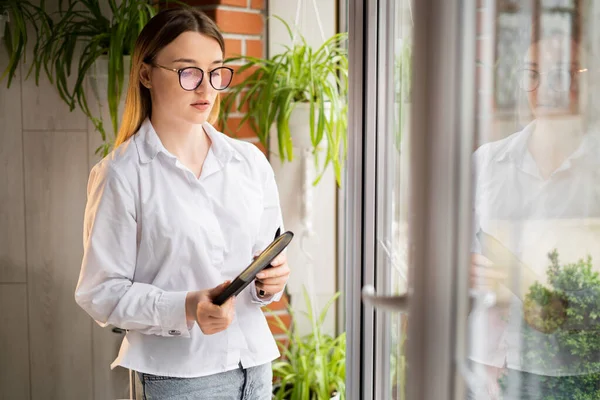 business woman standing in the office by the window with a notebook in her hands. Serious upset secretary with glasses at the panoramic window. Office worker with a paper client base. Work concept.