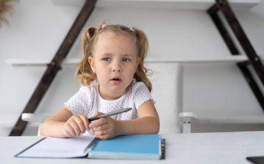 El yazısı çizmek için elinde defter ve kalemle masasında otururken evde ödevini yapan küçük tatlı bir ilkokul çocuğu. Öğrenci okulu uzaktan öğrenim görüyor. Anaokulu öğrencileri yazmayı öğrenirler.