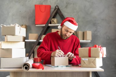 İş kurma KOBİ kavramı. Girişimci olmak isteyen genç bir erkek, küçük işletme sahibi, adresi kutunun üzerine yazıyor. Noel mevsiminde Noel Baba şapkası giyip satılıktı. Boşluğu kopyala.