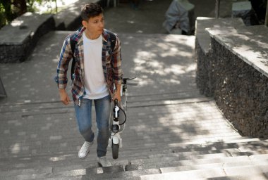Şehirdeki merdivenlerden katlanmış elektrikli scooterla çıkan bir adam. Ekolojik teknolojik yaşam tarzı. Elektronik hareketlilik. Katlanmış bir pozisyonda elektrikli scooter taşıyan bir adam. Eko Şehir Taşımacılığı. Boşluğu kopyala.