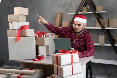 Evde ufak tefek eşyalar paketliyoruz. Noel Baba şapkalı bir adam Noel arifesinde ya da yeni yıl arifesinde masada otururken hediyelerle kutuları sayar. Ofis sahibi. Tatil teslimatı kavramı