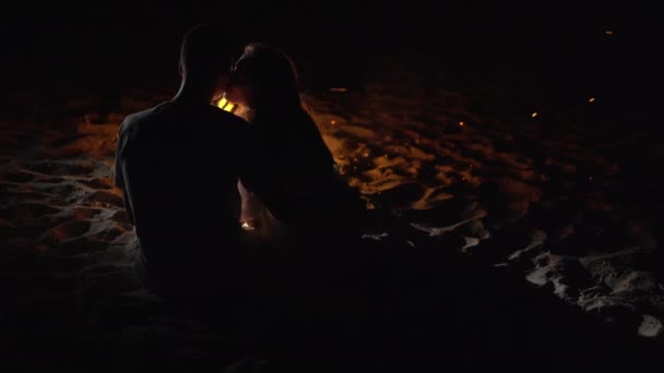 Kavkazský pár muž a žena sedí večer na pláži u ohně a líbají. — Stock video