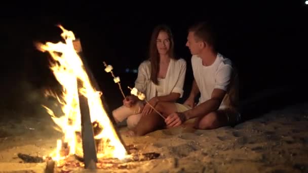 Egy fiatal pár pihen a tűz mellett, ül a homokos parton és mályvacukrot süt.. — Stock videók