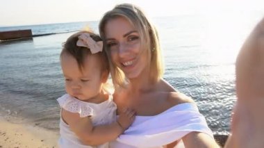 Günbatımında kumlu plajda görüntülü konuşma yapan mutlu anne ve kız. Anneler Günü. Küçük sarışın kız ve yurtdışında güzel turizm online video görüşmeleri..