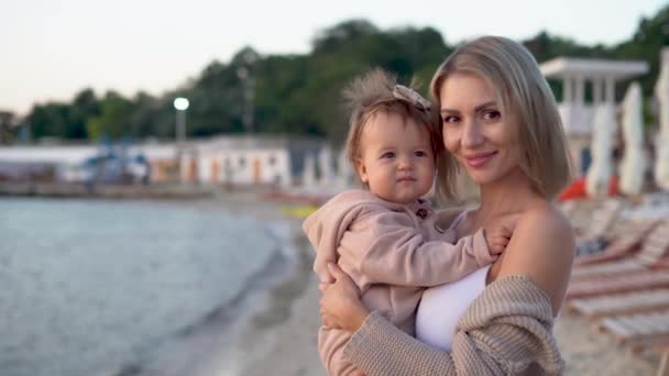 Zbliżenie portret mamy trzymającej córkę w ramionach podczas relaksu na plaży jesienią lub wiosną. Przestrzeń kopiowania. — Wideo stockowe