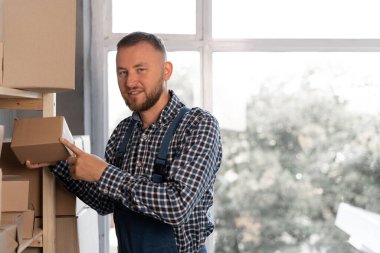 Mavi tulumlu, beyaz sakallı bir adam depodaki bir rafa bir kutu dolusu mal koyar. Boşluğu kopyala.