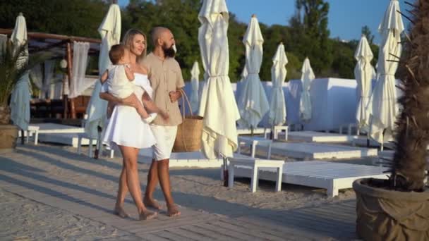 Caucasiano mãe família pai barbudo e pequena filha pré-escolar caminhar ao longo da praia entre espreguiçadeiras e palmeiras. — Vídeo de Stock