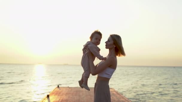 Mladá blondýna stojí ráno na molu, drží v náručí kojence a líbá svou dceru. — Stock video