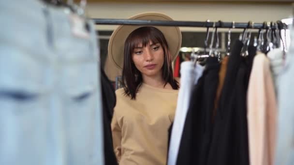 Mooie vrouw met een hoed kiest kleren in een kledingwinkel. Portret van een mooie lachende dame die iets te koop vindt in een kledingwinkel. — Stockvideo