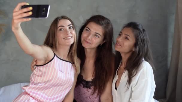 Three cheerful women friends in pajamas, sitting on the bed at home, take a selfie. — Stock Video