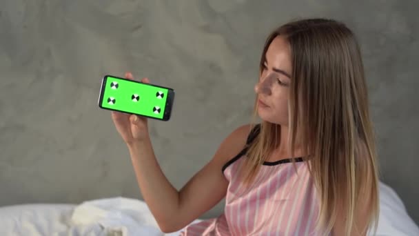 Femme caucasienne souriante des points milléniaux au smartphone moderne et pointant avec l'index à la maquette téléphone portable avec écran vert. Technologie, gadget, concept publicitaire. — Video