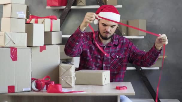 Homem caucasiano pequeno empresário senta-se em um escritório em casa usando um chapéu de Papai Noel e amarrando um arco vermelho em uma caixa. conceito de venda de natal. — Vídeo de Stock