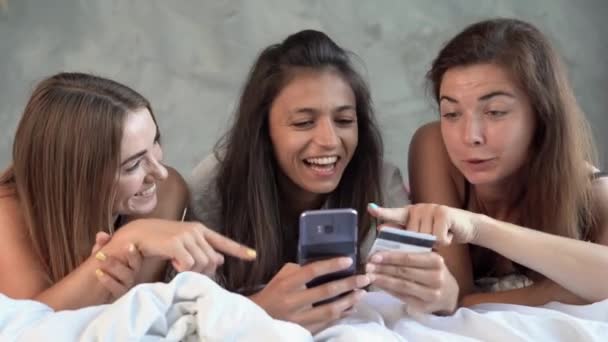 3人の魅力的な白人女性の友人は、午前中に自宅でオンラインショッピングを楽しんでいます。女性は、電子銀行を通じてオンライン決済のためのクレジットカードで寝室に横たわっています. — ストック動画