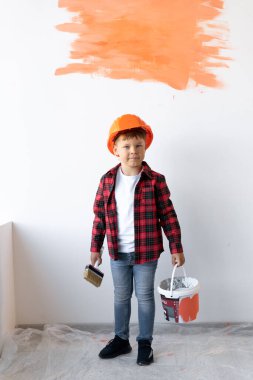 Gömlek ve kot giyen bir çocuk, elinde fırça ve boyayla turuncu kasklı beyaz bir duvarın yanında duruyor. Çocuk duvarı boyadı. Kendi evinizi kendiniz tamir edin. İnşaat alanı kavramı