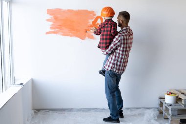 Evde tamirat yapan bir ailenin arkası görülüyor. Babası oğlunu kollarında tutuyor ve duvarı turuncuya boyuyor. Küçük bir ressamın kafasında bir kask. Evinin tadilatı. Dekoratör. Metin için yer