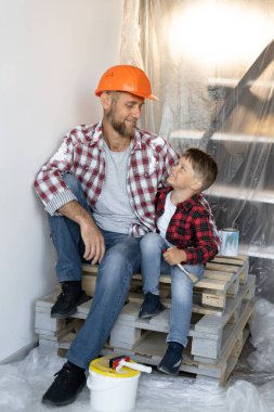 Evin restorasyonu, dekorasyonu ve boyanması, baba ve oğlu evin tadilatı sırasında oturup ara veriyor. Ebeveynlik yardım kavramı. Boşluğu kopyala.