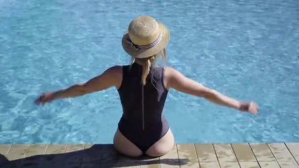 Back view of a beautiful young Caucasian lady sitting on the edge of the pool wearing a hat and a swimsuit enjoying her vacation on a tropical beach at a hotel. — Stock Video