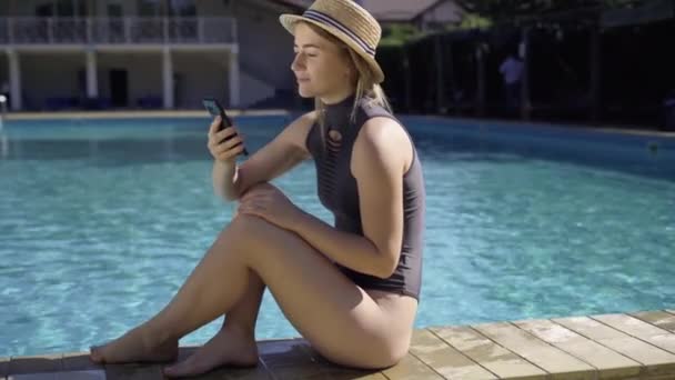 Young caucasian woman sitting near the pool leafs through the news feed on her smartphone. — Stock Video