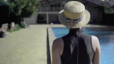 Arka planda, havuz kenarında hasır şapkalı, yaz tatilinin tadını çıkaran bir kadın var..