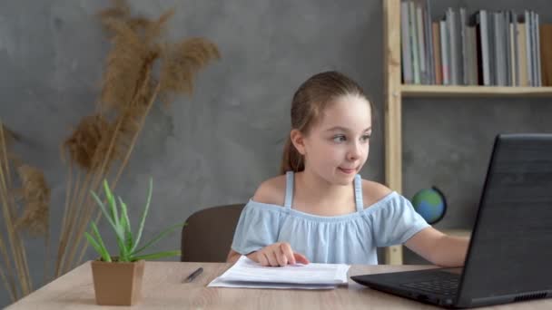 Colegiala preescolar inteligente haciendo la tarea con el ordenador portátil en casa. toma notas con un bolígrafo en un pedazo de papel. — Vídeos de Stock