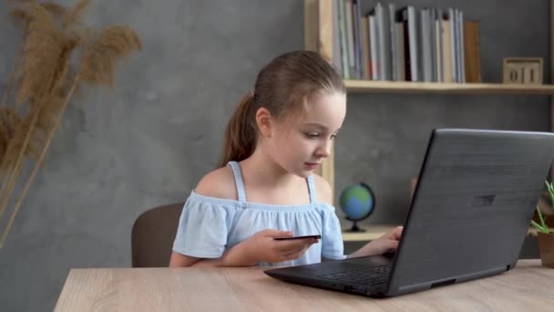 Alegre niña hace compras en línea utilizando un ordenador portátil y una tarjeta de crédito mientras se relaja en la mesa en casa. — Vídeos de Stock