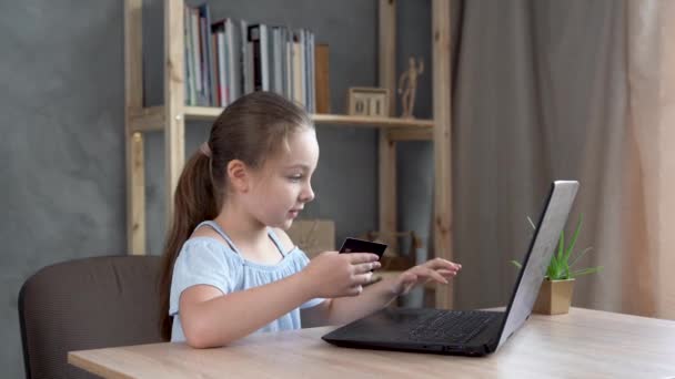 Alegre niña hace compras en línea utilizando un ordenador portátil y una tarjeta de crédito mientras se relaja en la mesa en casa. — Vídeos de Stock