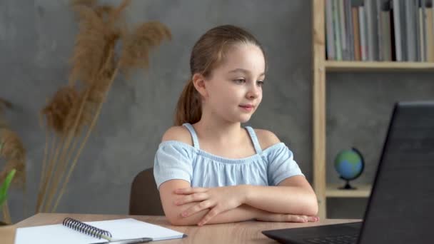 Una estudiante de primaria escucha una lección en línea a través de la comunicación por video en casa en su escritorio, estudiando de forma remota con una computadora portátil o PC. La colegiala está en contacto con un profesor en línea — Vídeos de Stock