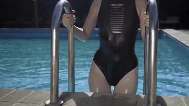Piękna młoda kobieta wychodzi z basenu, trzymając drabinę basenową. Uwodzicielska modelka w czarnym stroju kąpielowym przy basenie. Koncepcja odkrytego basenu. — Wideo stockowe