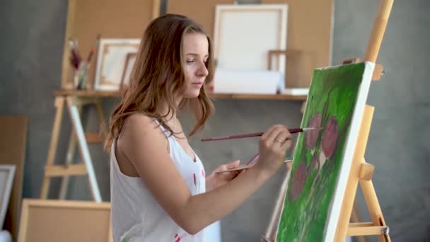 Portrait en gros plan d'une femme artiste elle peint un tableau avec des pivoines avec un pinceau et des peintures acryliques sur toile. Les instruments sont partout dans le studio. — Video